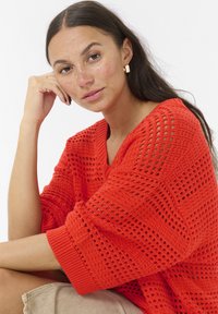 Pulover tricotat, de culoare portocalie, cu un design texturat și lejer, guler în V și mâneci largi, asortat cu pantaloni în culori neutre.