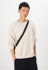 Felpa beige con scollo rotondo e maniche lunghe, caratterizzata da un sottile dettaglio di design sul petto. Pantaloni neri, stile minimale.