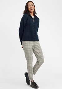 Navy cable-knit pullover with half zip, paired with light plaid tapered pants. Model wears black lace-up shoes. White background.