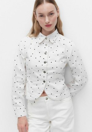 Vrouw die een wit jasje met zwarte polkadots en bijpassende witte broek draagt, staand tegen een effen witte achtergrond.