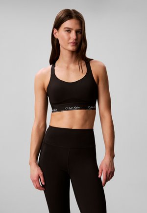 Femme portant un soutien-gorge de sport noir Calvin Klein et un legging taille haute assorti, debout devant un fond gris uni.