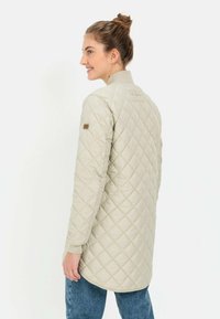 Hellbeige gesteppte Jacke mit langen Ärmeln, gerippten Bündchen und einem leicht verlängerten Design. Verfügt über ein kleines Logo-Patch auf der Schulter.