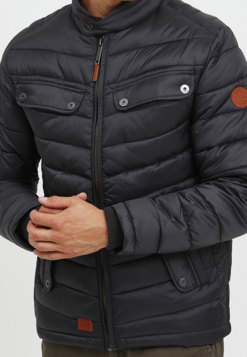 Veste matelassée noire avec un col montant, comprenant deux poches poitrine, une fermeture éclair et un patch en cuir marron. Texture lisse, design structuré.