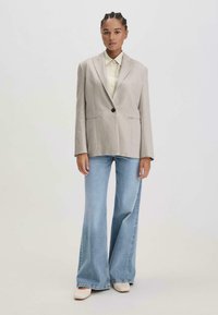 Beige maßgeschneiderter Blazer mit einem einzigen Knopf, kombiniert mit einem hellgelben Hemd mit Kragen und weit geschnittenen hellblauen Jeans. Weiße Schuhe runden das Outfit ab.