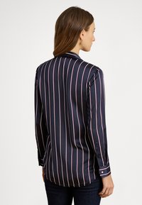 Sieviete ar brūniem matiem, valkājot tumši zilu blūzi ar garām piedurknēm un baltām un sarkanām vertikālām svītrām, skatās prom no kameras.