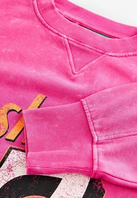 Sudadera rosa con cuello y puños acanalados, que presenta una impresión gráfica desgastada en amarillo y negro contrastante en la parte frontal.