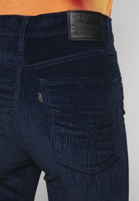 Calças de ganga de veludo cotelê azul-escuro com uma superfície texturizada, bolsos traseiros com costura decorativa e uma etiqueta de marca em couro preto na cintura.