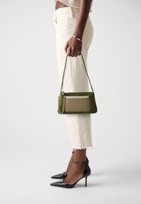 Borsa a tracolla verde in pelle con finitura testurizzata, accentata da dettagli crema e con tracolla a catena, tenuta da una persona che indossa pantaloni bianchi e tacchi neri.