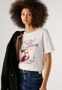 Maglietta di cotone bianco con stampa grafica con testo rosso e nero. Abbinata a denim blu scuro e cintura nera con borchie.