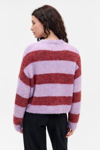 Donna con capelli ricci scuri che indossa un maglione peloso a righe lilla e rosse, girata di lato e che guarda verso sinistra su uno sfondo bianco.