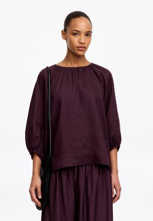 Femme portant un chemisier ample à manches longues violet foncé et une jupe assortie, portant un sac à bandoulière noir sur fond blanc.