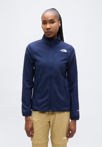 Γυναίκα που φοράει μπλε ζακέτα με φερμουάρ και μπεζ παντελόνι, στέκεται μπροστά σε απλό λευκό φόντο, κοιτάζοντας μπροστά με ουδέτερο ύφος.