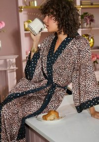 Accappatoio in stampa leopardata con dettagli blu navy e stelle dorate; tessuto leggero con maniche ampie, cinturato in vita. Tazza e cornetto su un tavolo.