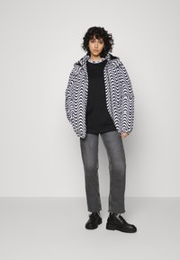 Casaco com padrão zigzag em preto e branco, com capuz, sobre uma blusa preta. Combinado com jeans cinza desgastados e botas pretas.