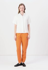 Camicetta bianca a maniche corte con taglio dritto, abbinata a pantaloni della tuta arancioni con un logo verde, e scarpe nere senza lacci.