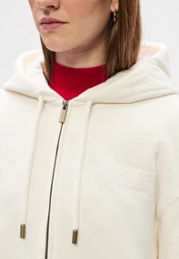 Cremfarbene Kapuzenjacke mit Reißverschluss, Fronttaschen und Kordelzügen. Verfügt über ein rotes geripptes Innenfutter und dezente bestickte Markenlogos auf der Brust.