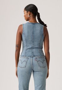 Γυναίκα με κοτσίδες, φοράει στενό αμάνικο τζιν τοπ και ανοιχτόχρωμο τζιν, στέκεται γυρισμένη με την πλάτη προς το απλό φόντο.