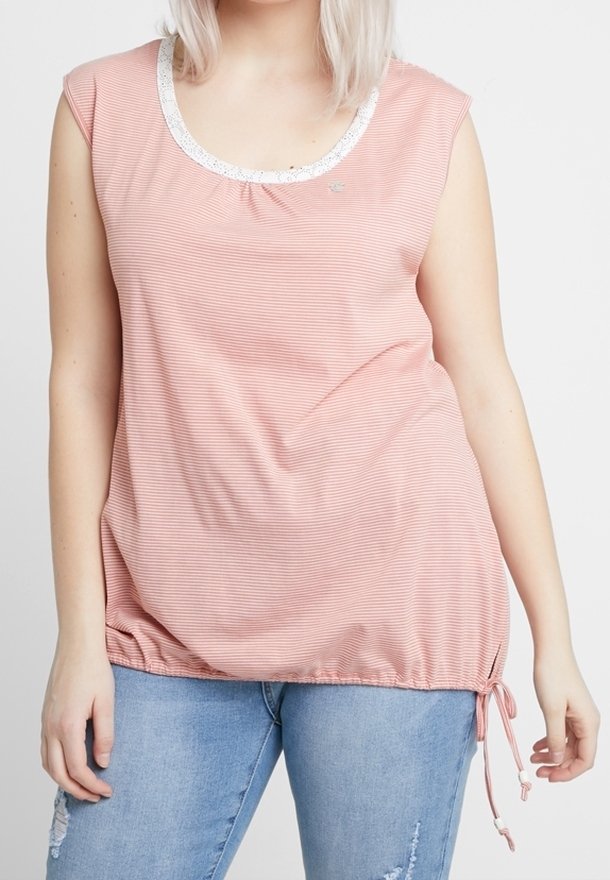 Femme portant un haut sans manches rose rayé avec un bord en dentelle blanche et un jean bleu clair déchiré, debout devant un fond uni.