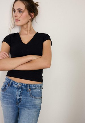 Giovane donna con le braccia incrociate che indossa una maglietta nera con scollo a V e jeans blu, che guarda a sinistra su uno sfondo chiaro semplice.