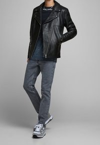 Svart läderbikerjacka med dragkedja fram och bred krage, kombinerad med en marinblå grafisk T-shirt, grå denimjeans och grå sneakers.