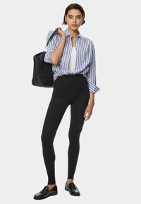 Svarta tights med hög midja, kombinerat med en blå och vit randig skjorta med knappar. Accessoarer inkluderar en texturerad svart tote-väska och svarta loafers.