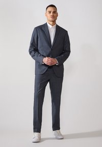 Completo grigio scuro con motivo a quadri, giacca monopetto, pantaloni abbinati, camicia bianca e sneakers bianche.