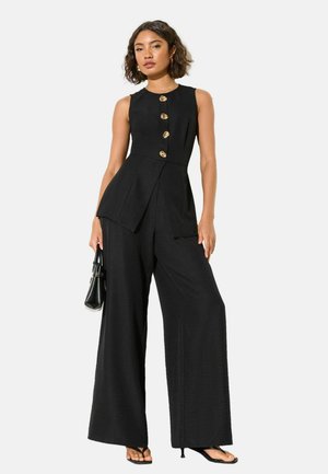 Frau trägt ärmellosen schwarzen Jumpsuit mit goldenen Knöpfen, weiten Hosenbeinen, hält eine schwarze Handtasche und trägt schwarze Sandaletten mit Absatz.