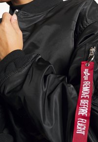Czarna kurtka bomberka o gładkiej fakturze, z prążkowanymi mankietami i czerwoną metką z napisem "USUNIĘĆ PRZED LOTEM" przypiętą do zamka.