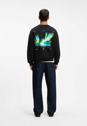 Homme portant un sweat-shirt noir avec un motif coloré d'aurores boréales dans le dos et un jean bleu foncé à jambes larges, debout de dos.