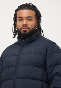 Mand med mellemlange dreadlocks og skæg iført en marineblå quiltet jakke med høj krave og lille logo på brystet.