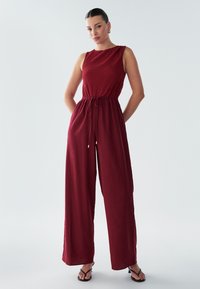 Burgundski brezrokavnik z teksturirano zgornjo stranjo, pasom z zavezovanjem in širokimi hlačnicami. Gladek material z minimalnimi detajli.