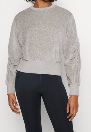 Woman wearing a light gray long-sleeve ruched sweatshirt with ribbed cuffs and hem, paired with black fitted leggings.