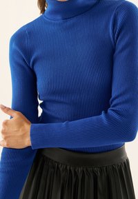 Femme portant un pull col roulé bleu à côtes et une jupe plissée noire, tenant doucement un bras avec une main.