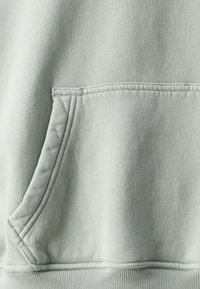Tejido de sudadera verde claro con una textura suave, que presenta un bolsillo tipo canguro en la parte frontal y detalles de costura a lo largo de los bordes.