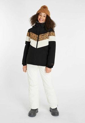 Giacca nera isolata con stampa leopardata e dettagli color crema. Pantaloni da neve bianchi e stivali neri. Cappello lavorato a maglia marrone con dettaglio a pom-pom.