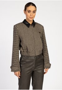 Frau mit nach hinten gekämmtem Haar, die eine graue Tweedjacke mit schwarzen, paillettenbesetzten Ärmeln und schwarzem Lederkragen trägt, kombiniert mit dunklen Lederhosen.