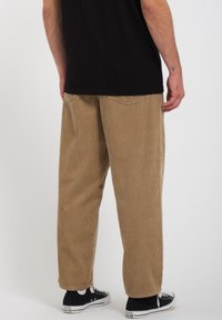 Ternet bukser i tan-farve med lodrette ribber, løs pasform og sidelommer. Båret med sorte sneakers med hvide detaljer.