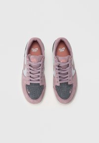 Par med Nike SB lavtop sneakers med rosa snøringer, grå tåkapper og rosa Nike swoosh på en hvit bakgrunn.