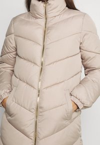 Chaqueta acolchada de un beige claro con costuras en forma de chevrón, cuello alto y cremallera dorada. Incluye bolsillos laterales y puños elásticos. Textura suave.