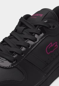 Baskets noires avec lacets noirs, détails brillants, logo crocodile rose sur le côté et marque Lacoste rose sur la languette.