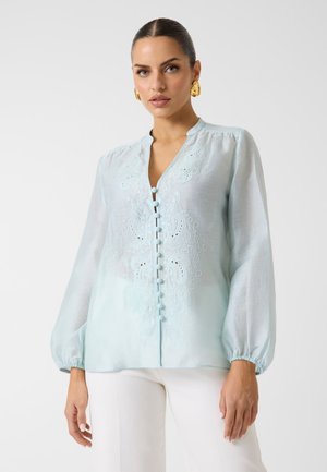 Frau mit zurückgestrichenen Haaren, die eine hellblaue bestickte Bluse mit Knöpfen vorne und weiße Hosen trägt, posiert vor schlichtem Hintergrund.