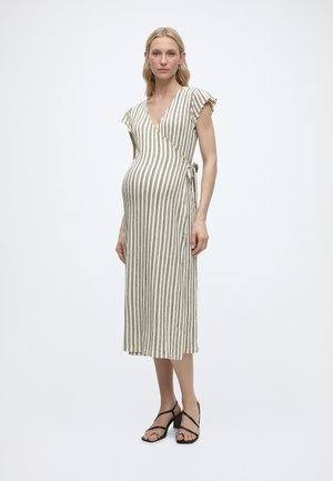 Zwangere vrouw staat, draagt een witte midi wikkeljurk met zwarte verticale strepen en zwarte bandjes blokhakken sandaal.