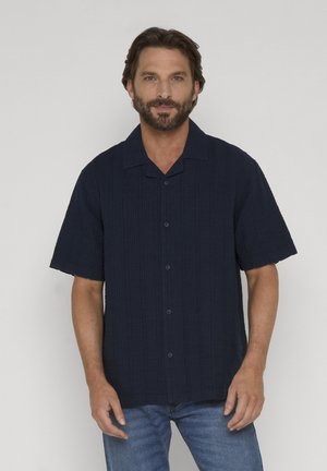 Homme barbu portant une chemise à manches courtes bleu foncé boutonnée et un jean bleu, debout devant un fond clair uni.