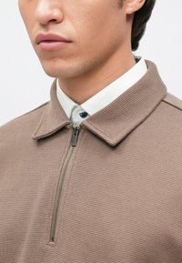 Pullover castanho com fecho éclair e gola, apresentando um tecido texturizado e uma camisa branca contrastante por baixo. O material do fecho éclair é visível.