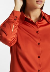 Blusa rossa di raso con colletto, maniche lunghe, polsini con bottone e dettagli ritagliati discreti sulle maniche. Finitura liscia e strutturata.