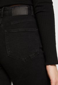 Los pantalones vaqueros de mezclilla negra presentan una cintura alta, una textura suave y un bolsillo trasero rectangular con costuras tonales. La tela tiene un diseño minimalista.