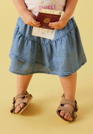 Child wearing sandals and a blue skirt holding a Danish passport and boarding pass against a plain background.