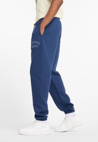 Person trägt blaue New Balance Jogginghosen und weiße Sneakers, steht mit einer Hand in der Tasche vor einem weißen Hintergrund.