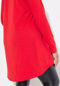 Rotes Langarm-Oberteil aus weichem, strukturiertem Stoff, mit lockerer Passform und abgerundetem Saum, kombiniert mit schwarzen, eleganten Leggings.