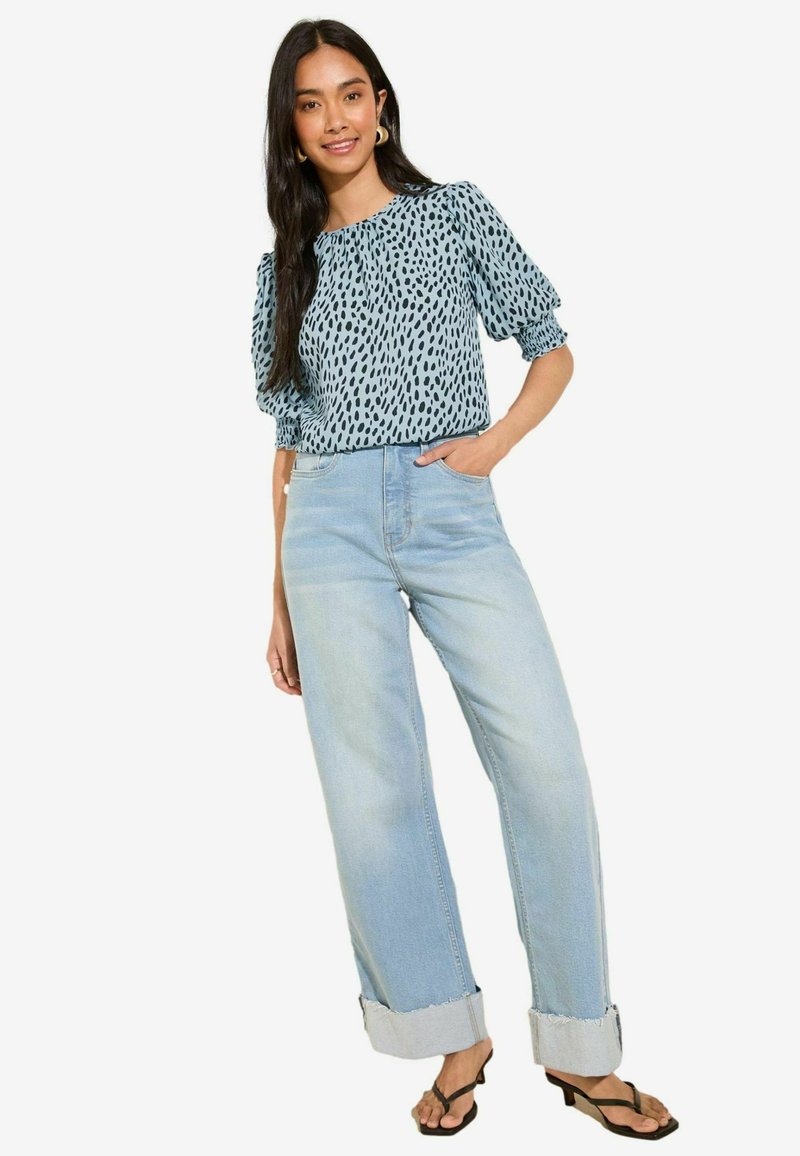 Blusa azzurro chiaro con motivo a pois neri, maniche a palloncino, abbinata a jeans a gamba larga di lavaggio chiaro, risvoltati all'orlo, e infradito nere.